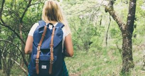 A girl with blonde hair wearing a white shirt and blue backpack standing in the wilderness 